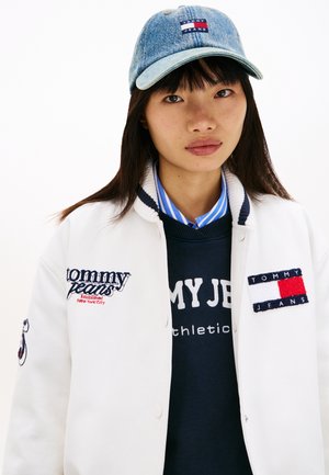 Hvid varsity-jakke med broderede logoer, navyblå sweater under med teksten "TOMMY JEANS" og blå denimkasket med logopatch.
