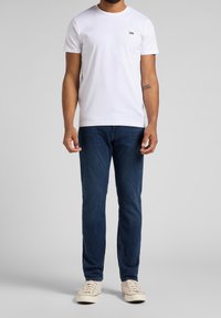 Hombre con camiseta blanca de Lee, jeans azul oscuro y zapatillas color crema, de pie sobre un fondo gris.