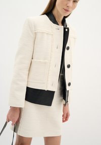 Giacca bianca testurizzata con colletto in pelle nera, caratterizzata da quattro bottoni e dettagli di cucitura visibili. Abbinata a una gonna testurizzata coordinata.