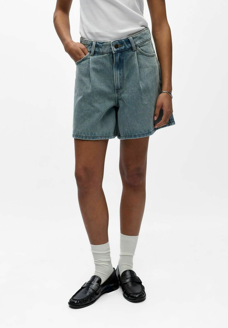 Personne portant un short en denim bleu clair, des chaussettes blanches, des mocassins noirs et un haut blanc sans manches sur un fond blanc uni.