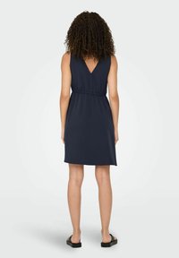 Marinefarbendes ärmelloses Kleid mit V-Ausschnitt hinten, tailliertem Schnitt und glattem Stoff, der über das Knie fällt, kombiniert mit schwarzen Sandalen.