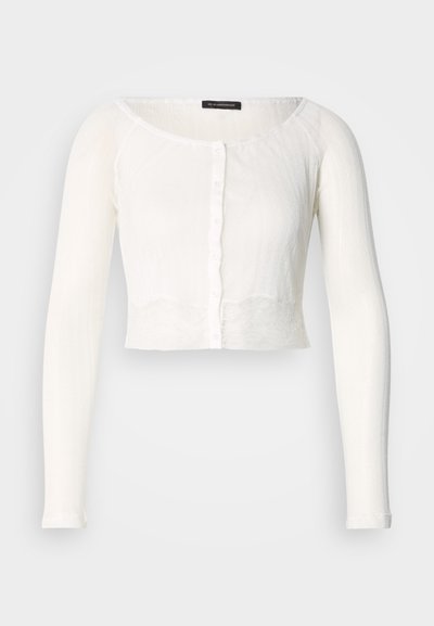 Λευκή κοντή ζακέτα με μακριά μανίκια, στρογγυλή λαιμόκοψη, υφασματική υφή και κούμπωμα με κουμπιά στο μπροστινό μέρος. Απαλό, ελαφρύ ύφασμα.