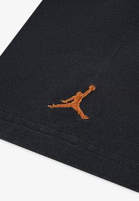 Juodas audinys su riešo tekstūra turi oranžinį siuvinėtą Jumpman logo. Dizainas demonstruoja sportinio drabužių estetiką.