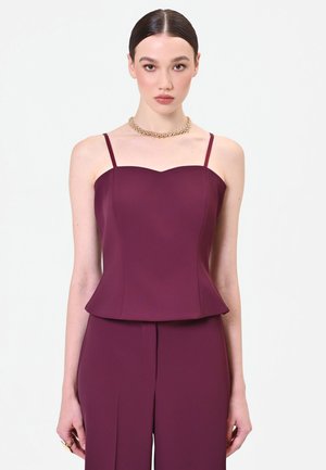 Giovane donna con capelli pettinati all’indietro indossa un top senza maniche color prugna e pantaloni abbinati, accessoriati con una collana dorata importante.