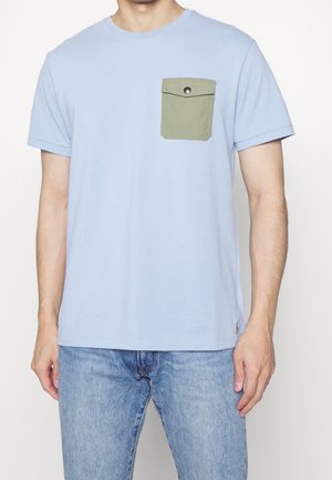 Mężczyzna w jasnoniebieskiej koszulce z krótkim rękawem, z oliwkowozieloną zapinaną kieszenią na piersi i jasnoniebieskich dżinsach, stojący na tle białego tła.