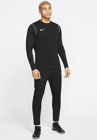 Zwarte sportieve top met lange mouwen en witte accenten, voorzien van het Nike-logo, gecombineerd met bijpassende zwarte tapered broeken en zwarte schoenen. Gladde textuur.
