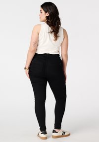 Vrouw met golvend haar, gekleed in een witte mouwloze top, zwarte high-waisted jeans, witte sneakers en armbanden, staand met haar rug naar de camera op een witte achtergrond.