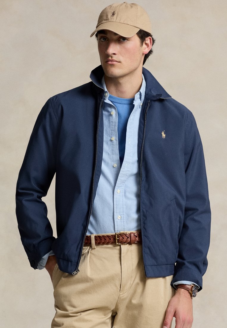 Veste zippée navy avec col montant, portée sur une chemise bleu clair à boutons. Une casquette beige, une ceinture en cuir tressé marron et un pantalon kaki complètent la tenue.