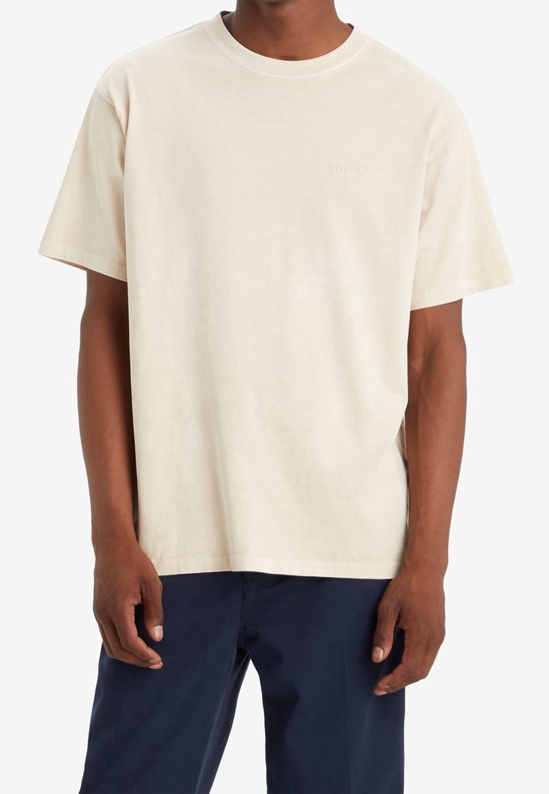 Homme portant un t-shirt beige uni à manches courtes et un pantalon bleu foncé, debout les mains détendues le long du corps.