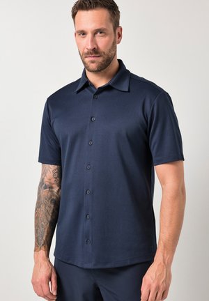 Man met kort haar en baard, draagt een donkerblauw overhemd met korte mouwen en knopen, toont zijn getatoeëerde linkerarm, staand tegen een effen achtergrond.