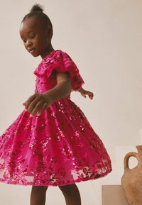 Skinnende pink kjole med blomsterformede pailletdetaljer og puffede ærmer. Nederdelen breder sig ud og skaber en voluminøs form.