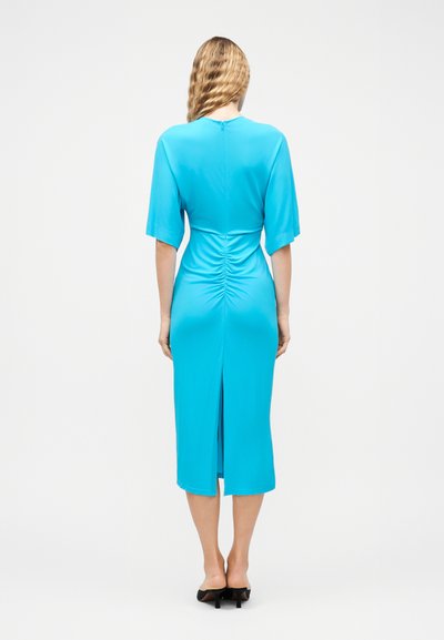 Γυναίκα με κυματιστά ξανθά μαλλιά φοράει εφαρμοστό, φωτεινό μπλε midi φόρεμα με πτυχές στην πλάτη και κεντρική σχισμή, συνδυασμένο με μαύρες γόβες, στέκεται με την πλάτη προς τα εμάς.