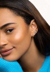 Pendientes en tono dorado con piedras preciosas azules y verdes; diseños de aro y de botón; llevados en la oreja derecha; sobre un suéter azul texturizado.