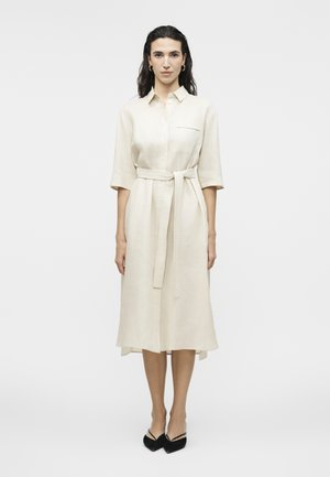 Γυναίκα που στέκεται με το πρόσωπο προς τα εμπρός, φορώντας ένα μπεζ φόρεμα με κουμπιά μέχρι το μέσο της γάμπας, με μανίκια τριών τετάρτων, ζώνη στη μέση που δένει και μαύρα παπούτσια με μυτερή μύτη.