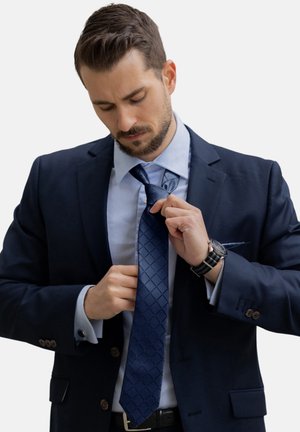 Marineblauer Anzug mit einem hellblauen Hemd und einer gemusterten marineblauen Krawatte. Die Person richtet die Krawatte, während eine Uhr am Handgelenk sichtbar ist.