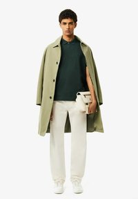 Olivgrön trenchcoat över en mörkgrön polo, vita byxor och vita sneakers. Modellen håller en liten krämfärgad crossbody-väska.