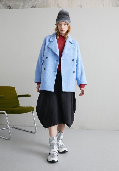 Manteau oversize bleu clair en mélange de laine, haut rouge, jupe noire et chaussures argentées avec poignets élastiques et détails en métal.