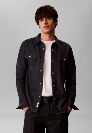 Junger Mann mit lockigem dunklem Haar trägt eine schwarze Jeansjacke über einem weißen Hemd und schwarzen Hosen mit den Händen in den Taschen vor einfarbigem grauem Hintergrund.