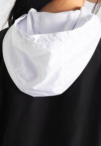 Černý svrchní oděv s bílou kapucí a strukturovanou síťovinou uvnitř. Kapuce má stáhnutý otvor pro nastavitelné padnutí.