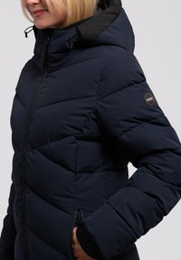 Giacca a vento blu navy imbottita con chiusura a zip, cappuccio e design trapuntato diagonale. Presenta una patch con logo sulla spalla e tasche laterali.