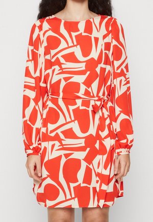 Mujer que lleva un vestido de manga larga con un patrón abstracto en rojo y blanco y un lazo en la cintura, de pie frente a un fondo liso.