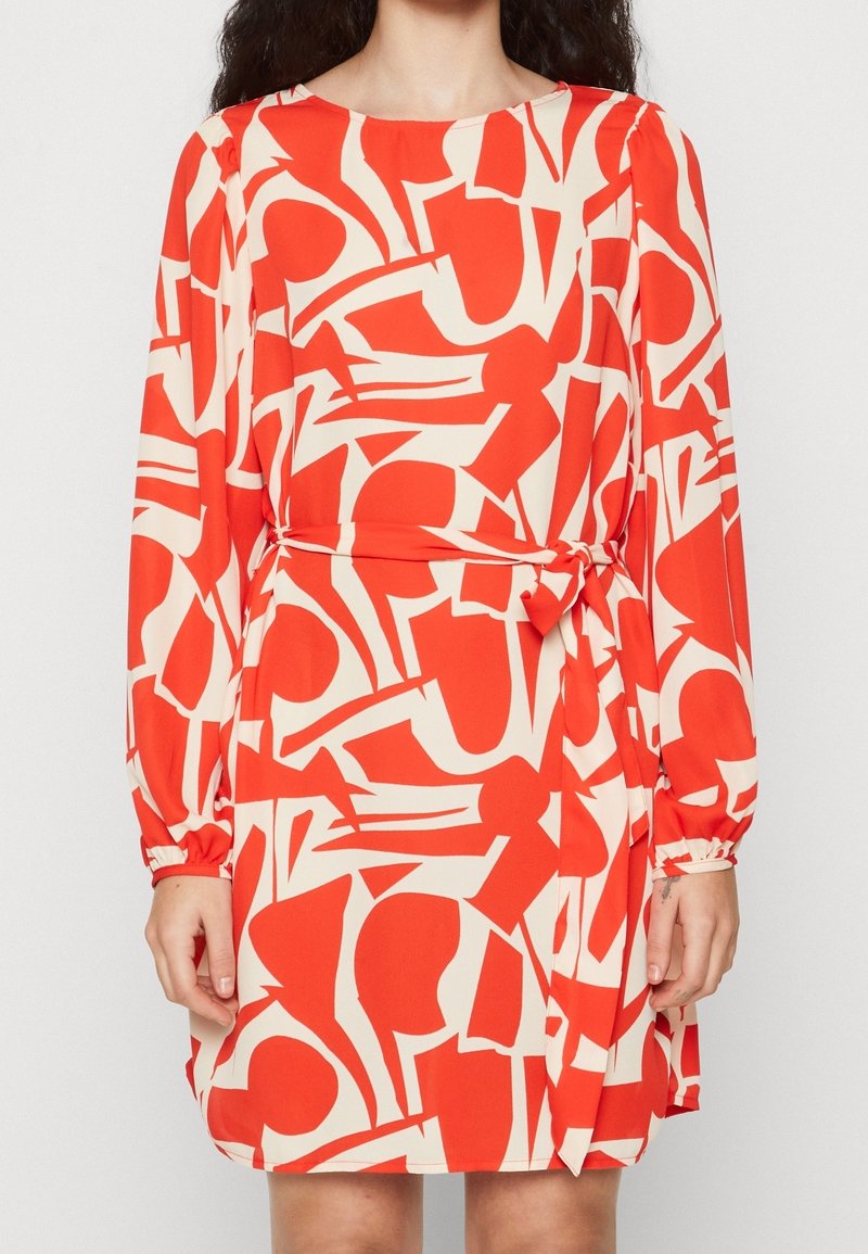 Femme portant une robe rouge et blanche à manches longues avec un motif abstrait et une ceinture à la taille, debout devant un fond uni.