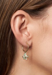 Pendiente de aro dorado con un pequeño colgante en forma de pez en verde y blanco. Presenta una textura suave y un diseño detallado. Se lleva en la oreja.