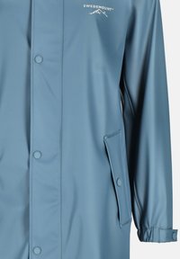 Niebieska wodoodporna kurtka przeciwdeszczowa z zapięciem na guziki z przodu, boczną kieszenią i regulowanymi mankietami. Lekka, gładka tekstura z detalem logo.