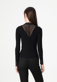 Donna che indossa un top a maniche lunghe nero con dettagli in pizzo sulla parte superiore della schiena e sulle maniche, abbinato a pantaloni neri aderenti, in piedi su uno sfondo bianco.