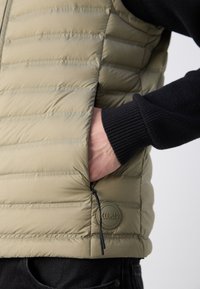 Gilet imbottito verde oliva chiaro in tessuto trapuntato. Presenta una tasca con zip e una toppa con logo rotondo. Maglione nero a maglia sotto.