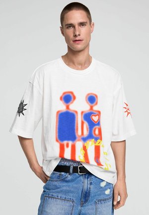 Mann trägt weißes oversized T-Shirt mit abstrakten blauen und roten Figuren, die Hände halten, schwarzer Sonnen-Grafik auf den Ärmeln und blauen Jeans.