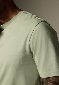 Svetlo zelena majica iz mehke tkanine z okroglim izrezom in kratkimi rokavi, z gladko teksturo brez vidnih vzorcev.