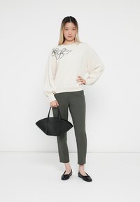 Lichtbeige sweatshirt met zwarte bloemenborduurwerk, gecombineerd met olijfgroene, aansluitende broek en een elegante zwarte handtas. Zwarte flats maken de outfit compleet.