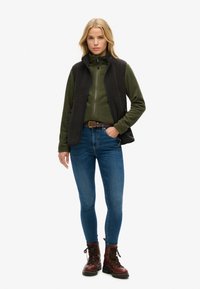 Zwarte fleece-vest over een groene jas met ritssluiting, gecombineerd met blauwe skinny jeans en bruine veterlaarzen. Eenvoudige, functionele buitenkleding.