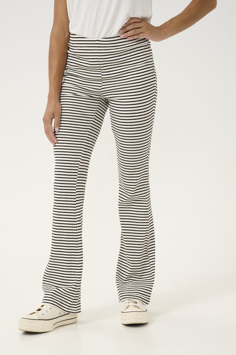 Femme portant un pantalon à rayures horizontales noires et blanches et des baskets blanches, debout, une main sur la hanche et l'autre détendue.