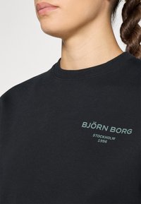 Fekete pulóver kerek nyakkal, puha anyagból készült, zöld nyomattal a mellkasán: "BJÖRN BORG STOCKHOLM 1956".