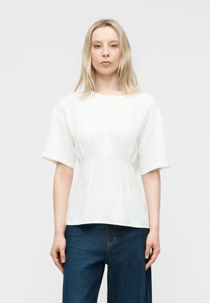 Ξανθιά γυναίκα που φοράει μια λευκή κοντομάνικη μπλούζα με υφή και μπλε τζιν, στέκεται μπροστά σε ένα απλό ανοιχτό γκρι φόντο.