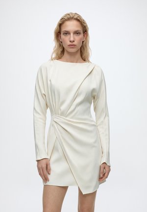 Un mannequin blonde se tient devant un fond uni clair, vêtue d’une mini-robe crème à manches longues et à jupe drapée asymétrique.