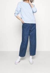 Sudadera de color azul claro con mangas largas y dobladillo acanalado, combinada con jeans de mezclilla oscuros de pierna ancha y cintura alta, y zapatillas blancas.