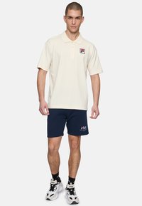 Camisa polo crema con cuello y un pequeño logo en negro, rojo y blanco; pantalones cortos azul marino; y zapatillas en blanco y negro con acentos contrastantes.