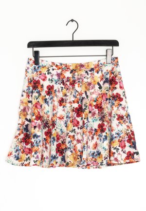 Plisowana spódnica w kwiaty w kolorach czerwonym, żółtym, niebieskim i różowym na białym tle, zawieszona na czarnym wieszaku z klipsem.