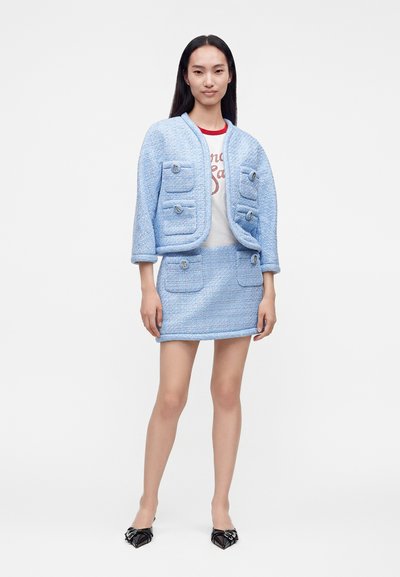 Femme portant un tailleur jupe bleu clair texturé avec quatre poches avant et une chemise blanche, debout avec des chaussures noires à bout pointu sur fond blanc.