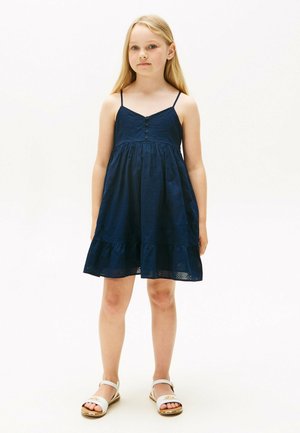 Marineblaue ärmellose Kleid mit Knopfdetails, Ösengewebe, ausgestelltem Rock und Rüschensaum; kombiniert mit weißen Sandalen.