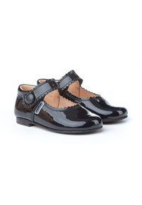 Zapatos de charol negro con punta redondeada, bordes festoneados y una correa en el tobillo con un botón redondo. Suela plana de goma.