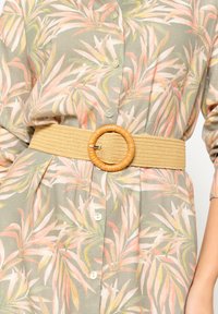 Cintura intrecciata beige con una fibbia rotonda in legno naturale testurizzato. La camicia presenta un motivo di foglie verdi e pastello con bottoni bianchi.