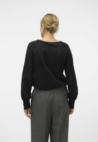 Kvinna med blont hår i en knut, klädd i en lös svart långärmad tröja och grå byxor med hög midja, som står mot en ren vit bakgrund.