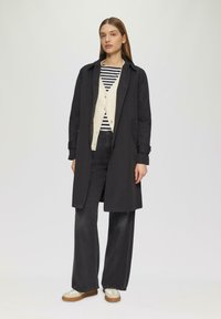 Zwarte trenchcoat over een crèmekleurige cardigan en gestreept shirt, gecombineerd met donkere wijde jeans en lichte sneakers. Vloeiende stof, open silhouet.