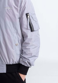 Bomber jacket grigio chiaro con polsini elastici, tasche laterali e tasca con zip sulla manica. Texture liscia e design casual.