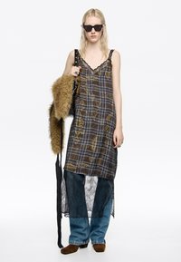 Vestido marrón a cuadros con encaje, superpuesto sobre encaje negro. Combinado con jeans de denim azul y zapatos de ante marrones. Bolso negro y gafas de sol.