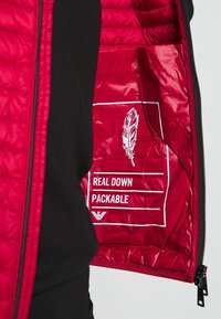 Červená péřová bunda s prošívaným designem, která ukazuje hladkou, lesklou podšívku. Obsahuje štítky "PRAVÉ PEŘÍ" a "SBALITELNÁ"; zapínání na zip.
